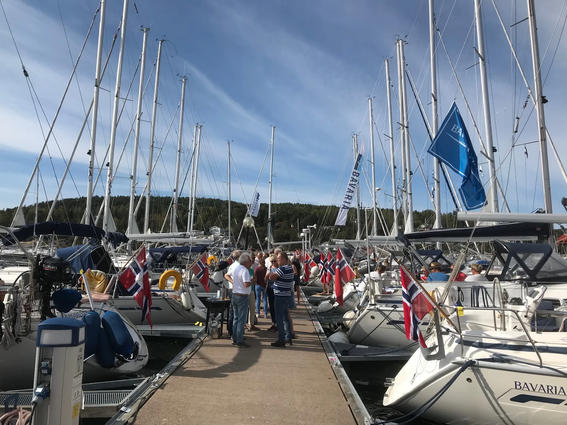 Medlemmer i prat på bryggen mellom Bavaria-seilbåter med norske flagg og Bavaria-vimpler under et klubbtreff